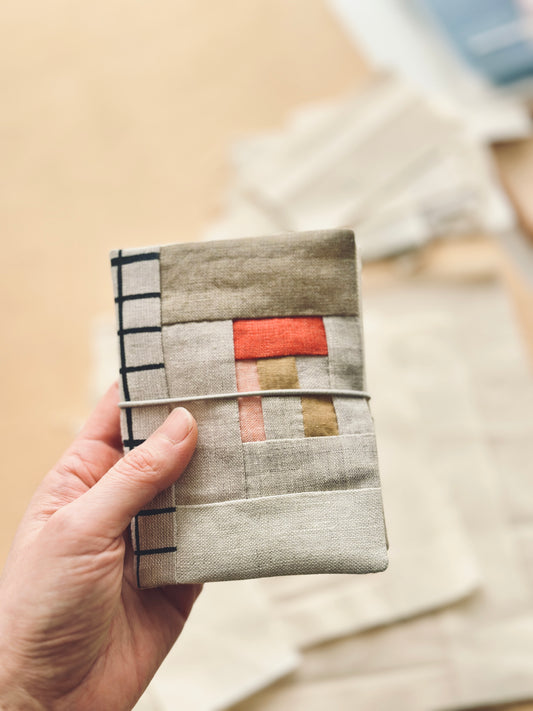 a hand holding a patchwork style linen needle book with crimson, soft pink, camel colour accent and black grid line fabric as spine with grey elastic closure