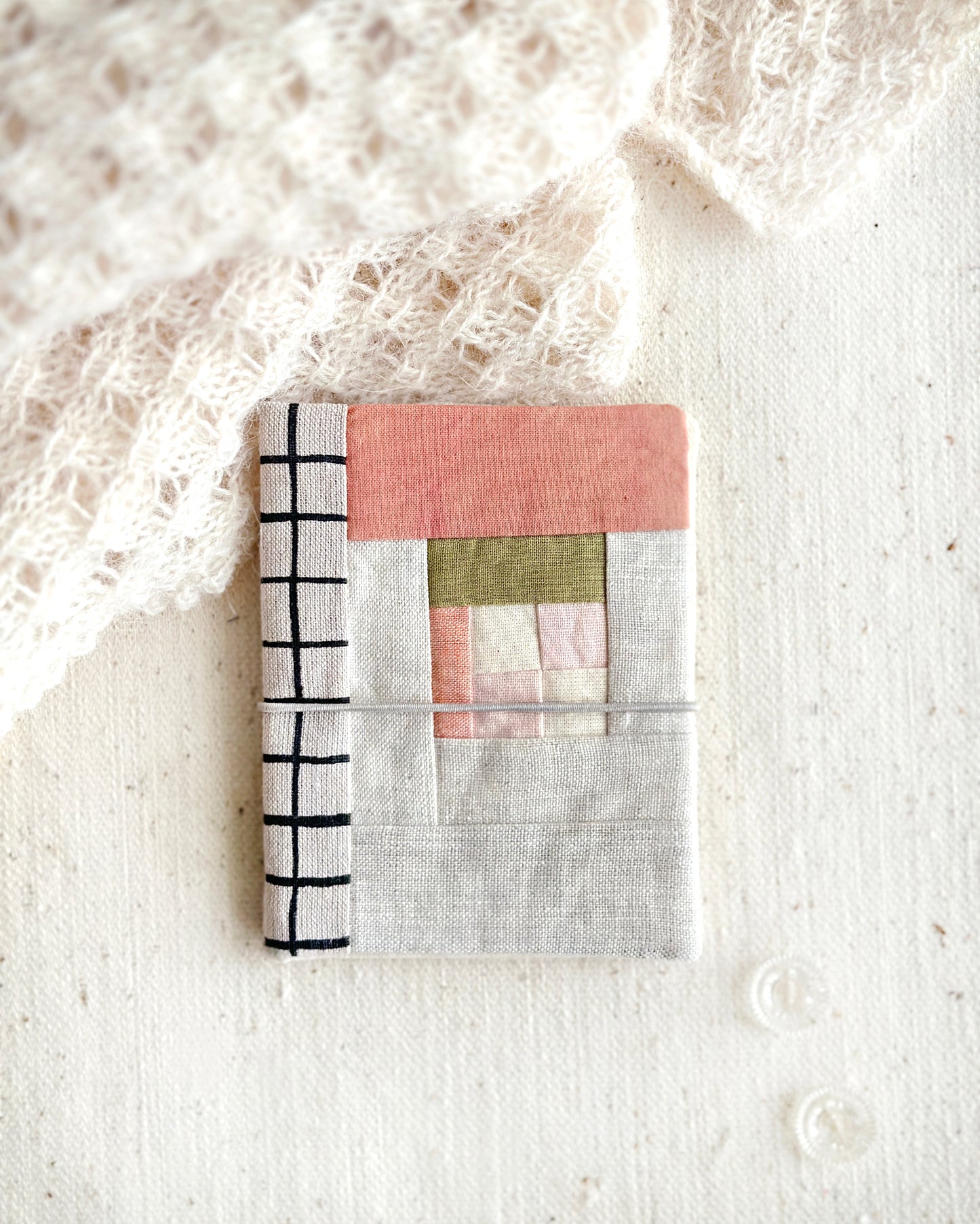 A patchwork needle case in soft peach, pink, and green tones combined with natural linen and black line grid spine laying on a textured cotton fabric with an off white lace scarf and two clear glass buttons