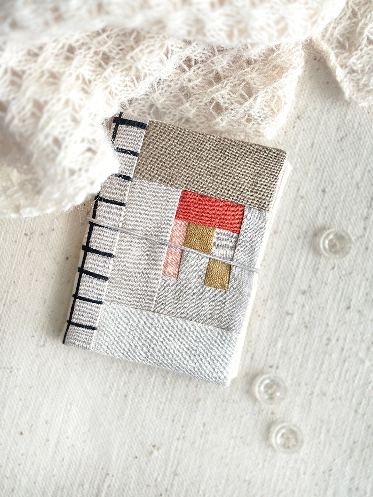 A needle case in red, soft pink,  and tan fabric patchwork combined with natural linen and black line grid spine laying on a textured white fabric with three clear glass buttons and beige lace scarf