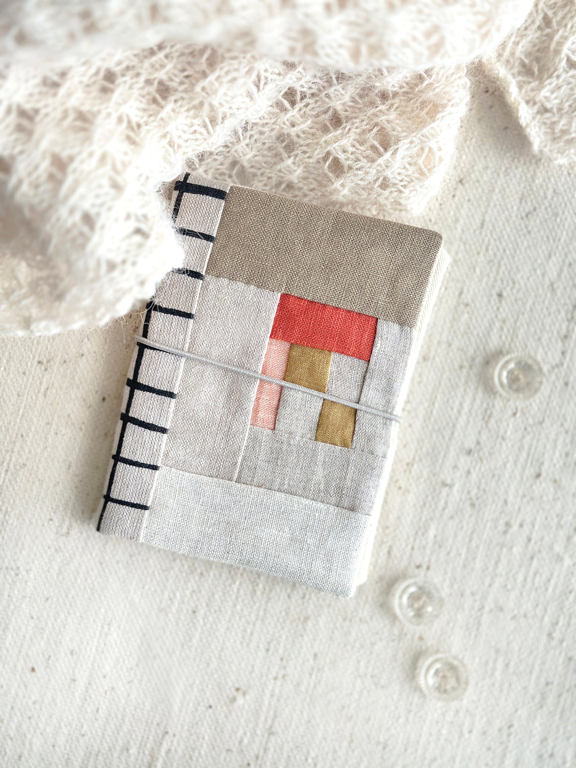 A needle case in red, soft pink,  and tan fabric patchwork combined with natural linen and black line grid spine laying on a textured white fabric with three clear glass buttons and beige lace scarf