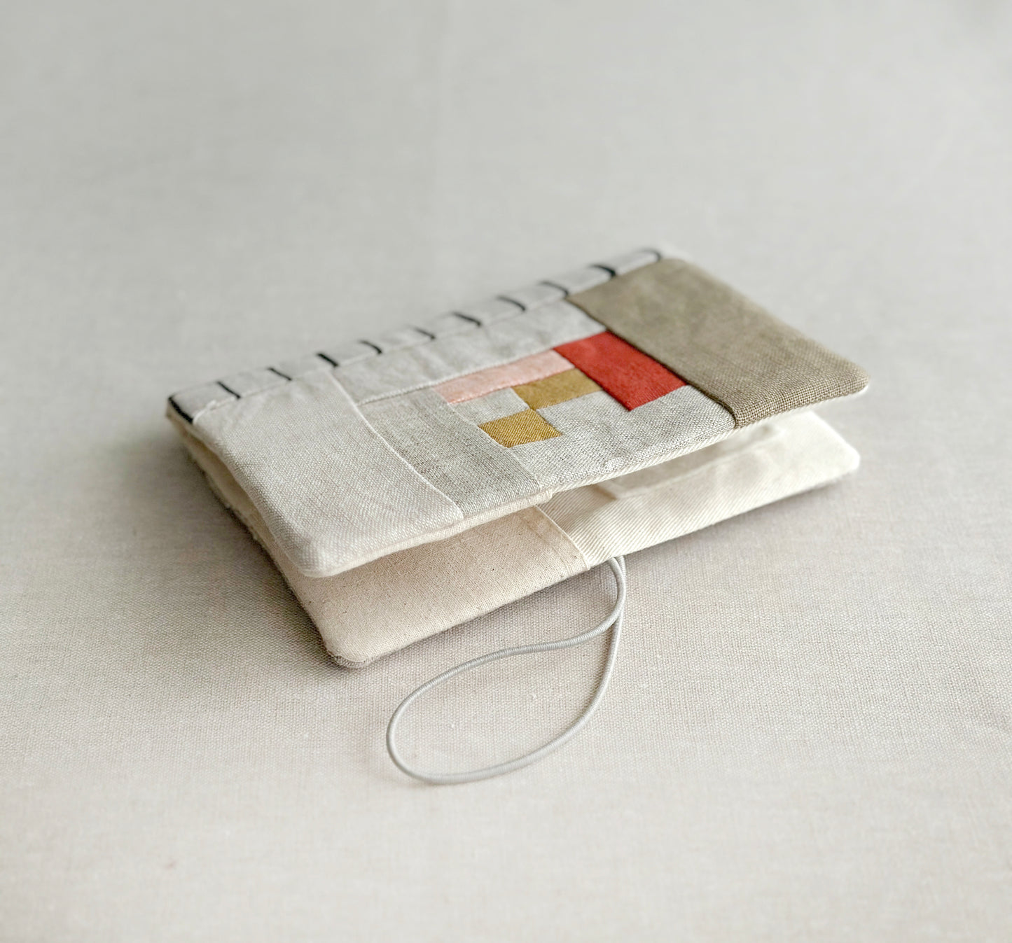 A needle case in red, soft pink,  and tan patchwork combined with natural linen and black lined pattern spine laying on a cream cotton fabric with elastic closure