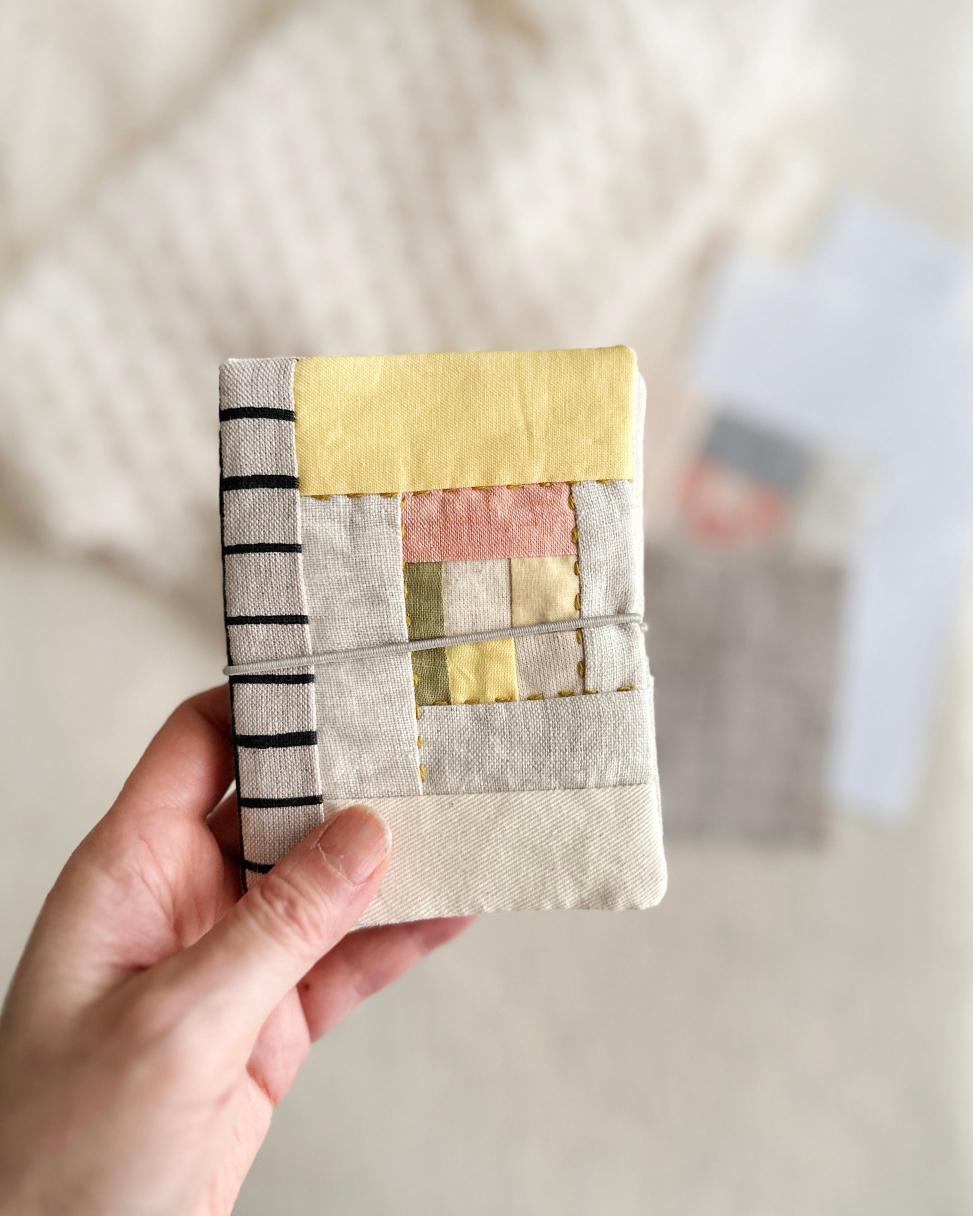 A hand held needle case in light yellow, soft pink and green fabric patchwork combined with natural linen and black line grid spine